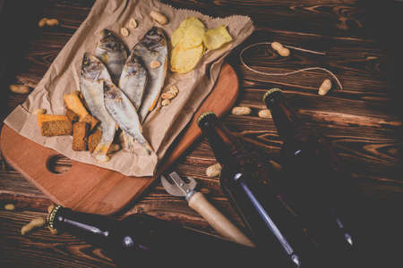 Brown glass bottles of beer and dried fish with chips, nuts, crackers on paper on a wooden background. Studio photo.の写真素材
