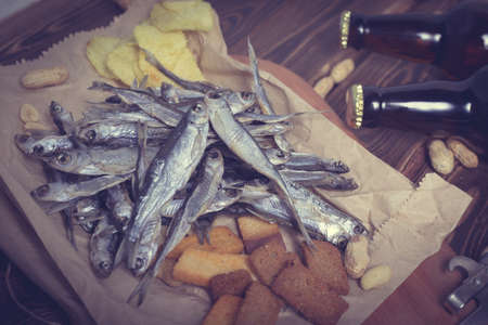 Brown glass bottles of beer and dried fish with chips, nuts, crackers on paper on a wooden background. Studio photo.の写真素材