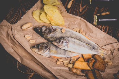 Brown glass bottles of beer and dried fish with chips, nuts, crackers on paper on a wooden...