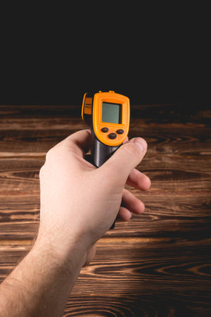A man holds a yellow-black pyrometer on a wooden background. A device for non-contact temperature...