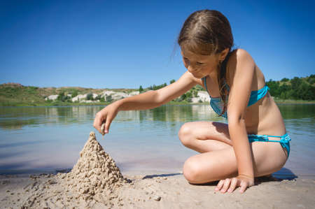 A little cute girl in a swimsuit is building a sand castle on the shore of a blue quarry lake.の写真素材