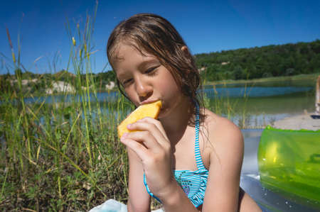 A little cute girl is eating her breakfast on the shore of a blue quarry lake.の写真素材