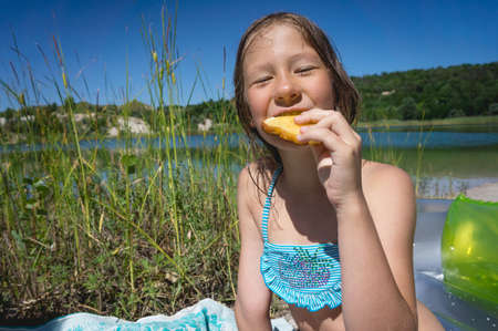 A little cute girl is eating her breakfast on the shore of a blue quarry lake.の写真素材