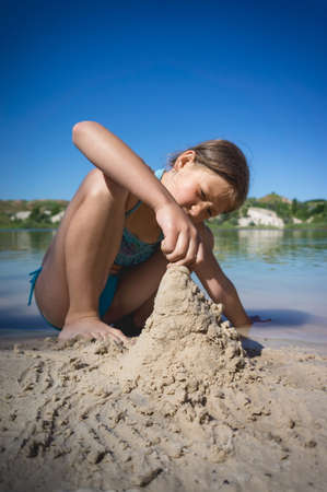 A little cute girl in a swimsuit is building a sand castle on the shore of a blue quarry lake.の写真素材
