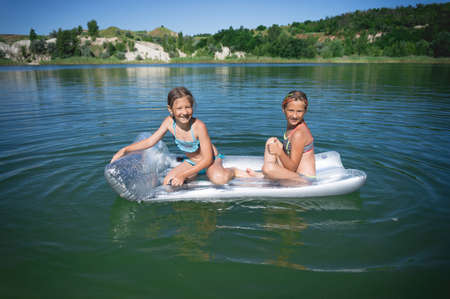 Two little cute girls in swimsuits swim on an inflatable mattress on a blue career lake.の写真素材