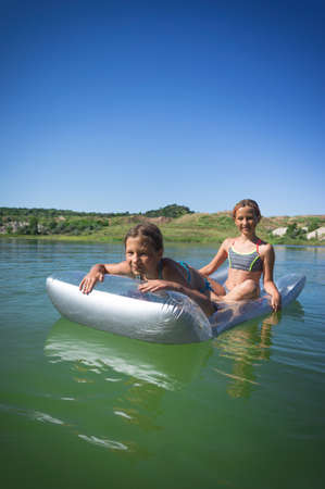 Two little cute girls in swimsuits swim on an inflatable mattress on a blue career lake.の写真素材