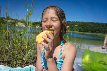 A little cute girl is eating her breakfast on the shore of a blue quarry lake.の写真素材