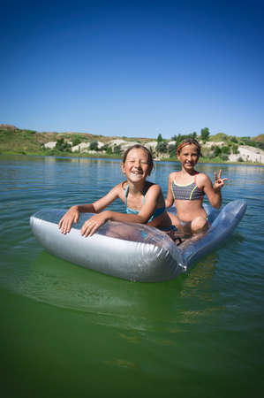 Two little cute girls in swimsuits swim on an inflatable mattress on a blue career lake.の写真素材