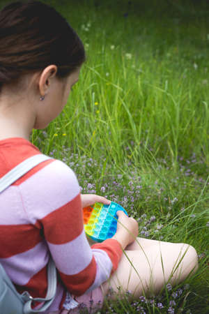 A little cute girl is sitting on the grass with a pop antistress toy.の写真素材