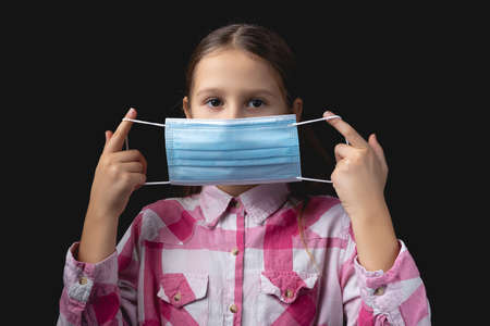 Little girl puts on a blue medical mask. Studio portrait isolated on black background with one light sourceの写真素材