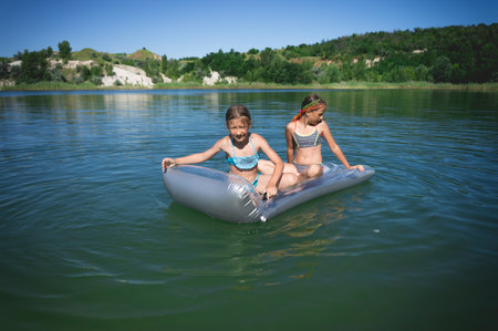 Two little cute girls in swimsuits swim on an inflatable mattress on a blue career lake.の写真素材