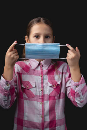 Little girl puts on a blue medical mask. Studio portrait isolated on black background with one light sourceの写真素材