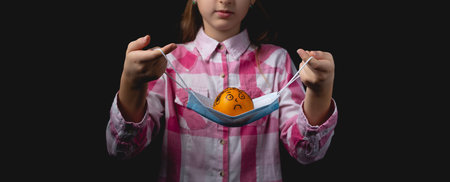 A little girl holds in front of her a blue medical mask with a tangerine in it. Mandarin painted with black felt-tip pen like a man without a mask. Studio portrait isolated on black background with one light sourceの写真素材