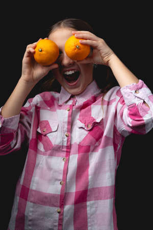 Little cute girl posing with tangerines in the studio. Photo isolated on black background with one light source. The child is fooling around closing his eyes with tangerines.の写真素材