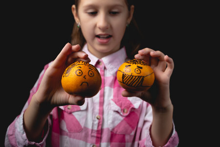 Little cute girl posing with tangerines in the studio. Photo isolated on black background with one light source. The child plays with tangerines painted like a man in a medical mask and a man without a mask.の写真素材