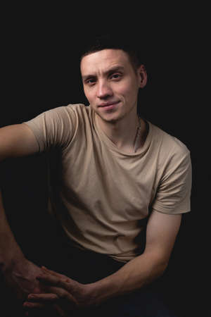 Handsome young man posing over black background. Studio photo with one light source. Smiling guy cheerfully posing in the studio. A serious guy sits with his elbow on his knee.の写真素材