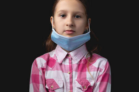 Little girl with a blue medical mask. The concept of wearing a medical mask incorrectly. Studio portrait isolated on black background with one light sourceの写真素材