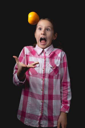 Little cute girl posing with tangerine in the studio. Photo isolated on black background with one light source. The child is fooling around by throwing a tangerine in the air in front of him.の写真素材