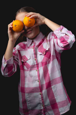 Little cute girl posing with tangerines in the studio. Photo isolated on black background with one light source. The child is fooling around closing his eyes with tangerines.の写真素材