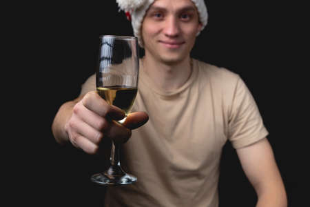Handsome young man with glass of champagne over isolated black background. A guy in a festive hat with a New Year's mood. Studio portrait with one light sourceの写真素材