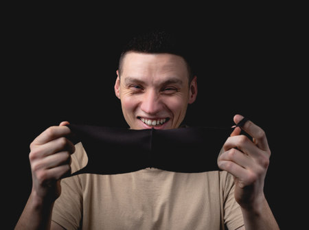 Handsome young man and a medical mask on a black background. The man is about to put on a medical mask. Studio photo with one light sourceの写真素材