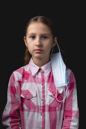 A little girl with a blue medical mask on one ear. Incorrect wearing of a medical mask. Studio portrait isolated on black background with one light sourceの写真素材