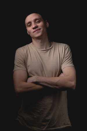 Handsome young man posing over black background. Studio photo with one light source. The confident guy crosses his arms in front of him and smiles.の写真素材