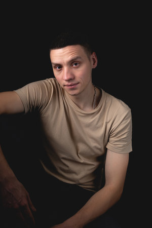 Handsome young man posing over black background. Studio photo with one light source. Smiling guy cheerfully posing in the studio. A serious guy sits with his elbow on his knee.の写真素材