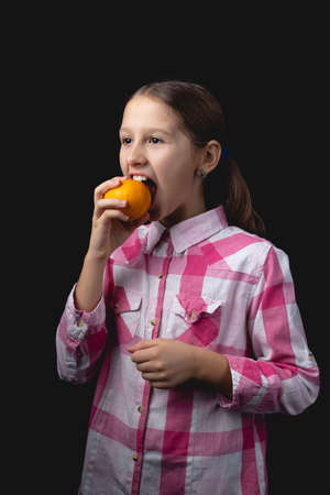 Little cute girl posing with tangerine in the studio. Photo isolated on black background with one light source. A child is fooling around trying to eat an unpeeled mandarin.の写真素材