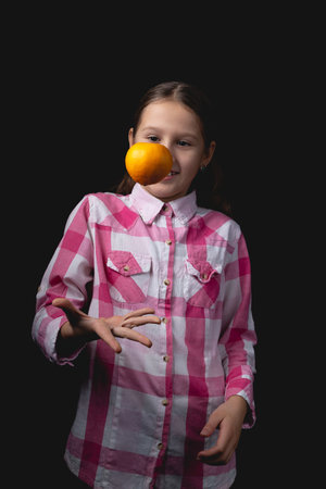 Little cute girl posing with tangerine in the studio. Photo isolated on black background with one light source. The child is fooling around by throwing a tangerine in the air in front of him.の写真素材