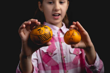 Little cute girl posing with tangerines in the studio. Photo isolated on black background with one light source. The child plays with tangerines painted like a man in a medical mask and a man without a mask.の写真素材
