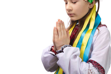 A little girl in national Ukrainian clothes with tearful eyes folded her hands in front of her and prays for an end to the war in Ukraine. Studio photo on a white background.の写真素材