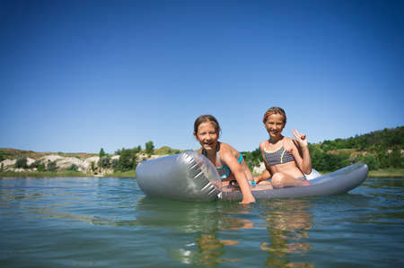 Two little cute girls in swimsuits swim on an inflatable mattress on a blue career lake.の写真素材