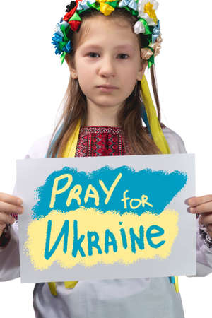 A little Ukrainian girl with tears in her eyes holds a sheet of paper with the inscription "Pray for Ukraine" in her hands. Studio photo on a white background.の写真素材