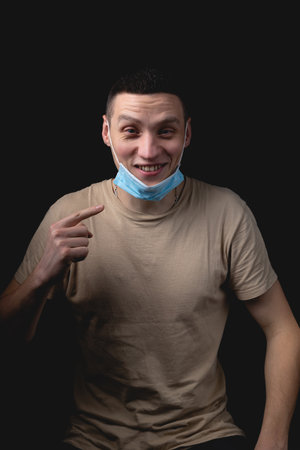 Handsome young man and a medical mask on a black background. The man shows how wrong it is to wear a medical mask.の写真素材