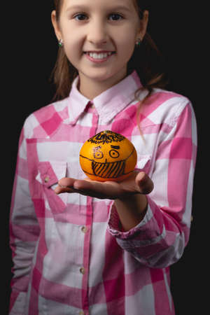 Little cute girl posing with tangerine in the studio. Photo isolated on black background with one light source. The child shows a tangerine painted like a man in a medical mask.の写真素材