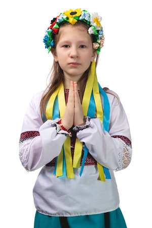 A little girl in national Ukrainian clothes with tearful eyes folded her hands in front of her and prays for an end to the war in Ukraine. Studio photo on a white background.の写真素材
