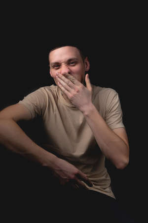 Handsome young man posing over black background. Studio photo with one light source. Smiling guy cheerfully posing in the studio. The guy covers his face with his hand, yawning.の写真素材