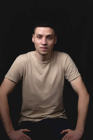 Handsome young man posing over black background. Studio photo with one light source. Smiling guy cheerfully posing in the studio. The guy sits leaning on his knees.の写真素材