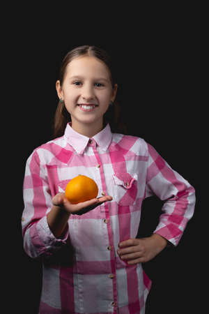 Little cute girl posing with tangerine in the studio. Photo isolated on black background with one light source. The child holds a tangerine in the palm of his hand.の写真素材