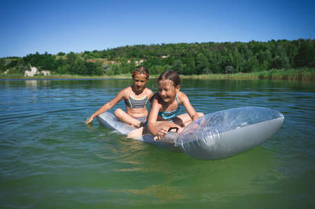 Two little cute girls in swimsuits swim on an inflatable mattress on a blue career lake.の写真素材