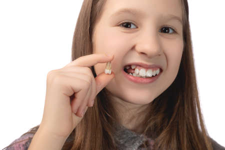A cute little girl holds a recently removed molar in her hand and shows the place from which this tooth was removed. Studio photo on a white background.の写真素材