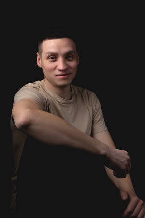 Handsome young man posing over black background. Studio photo with one light source. Smiling guy cheerfully posing in the studio. A cheerful guy sits with his elbow on his knee.の写真素材