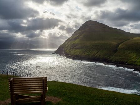 Faroe Islands. GjÃ³gv village. View form the hill above the village. Wooden bench in the foreground. The bay in the background. Dramatic sky with sun breaking through the clouds.の写真素材