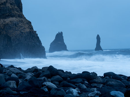 Reynisfjara Beach at dusk, with the iconic Reynisdrangar sea stacks standing against turbulent waves, framed by dark basalt columns and scattered volcanic rocksの写真素材
