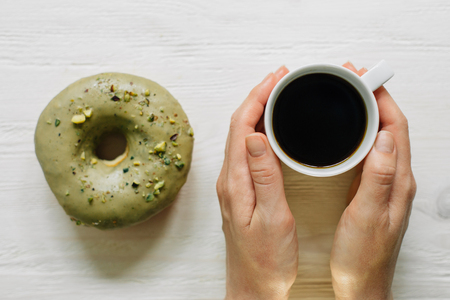 Girl's hands with cup of coffee and glazed almond donut behind. Light wooden backgroundの写真素材