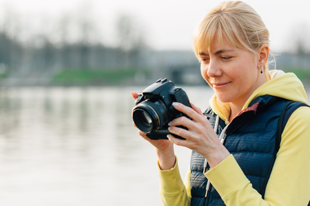 Girl with professional camera in hands. Close up of female photographer in park. Pretty smiling blonde woman looking at the screen of cameraの写真素材