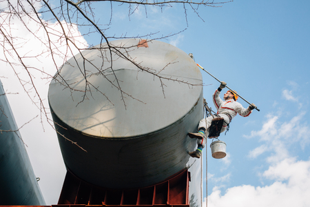 Industrial climber in helmet and uniform painting water tower. Professional Painter working on height. Risky job. Extreme occupation.の写真素材