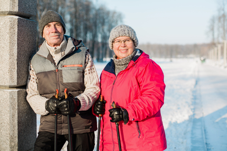 Pretty senior couple standing with nordic walking poles in winter park. Mature woman and old man resting outdoors. Healthy lifestyle concept.の写真素材