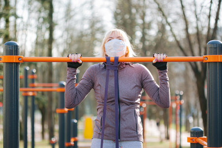 Woman in medical mask doing pull ups with rubber band. Training during coronavirus epidemic.